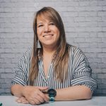 Portrait of a smiling woman wearing a striped shirt and watch, indoors against a brick wall.
